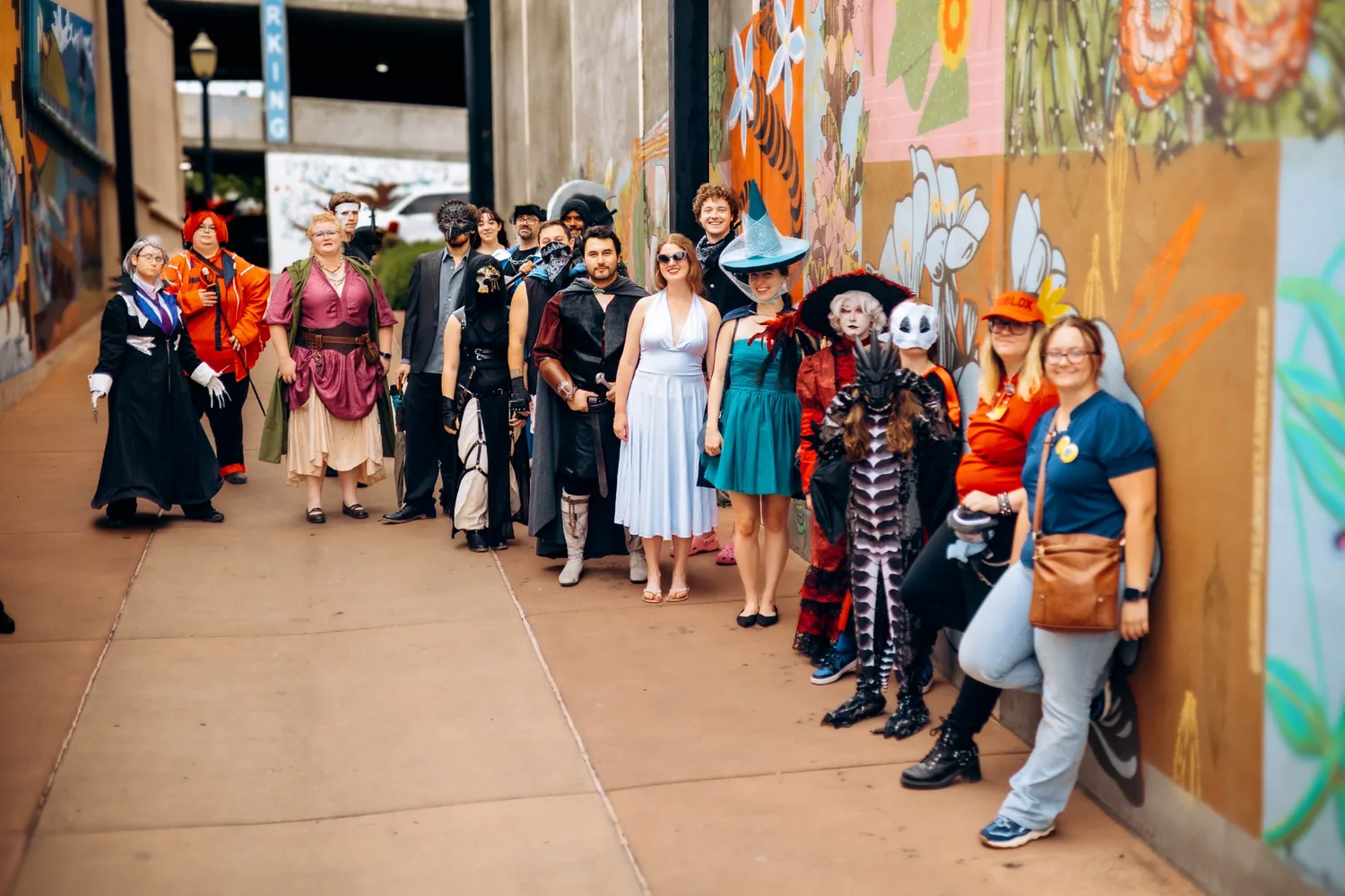 A large group in varied cosplay and everyday clothing gathered together beside colorful downtown murals