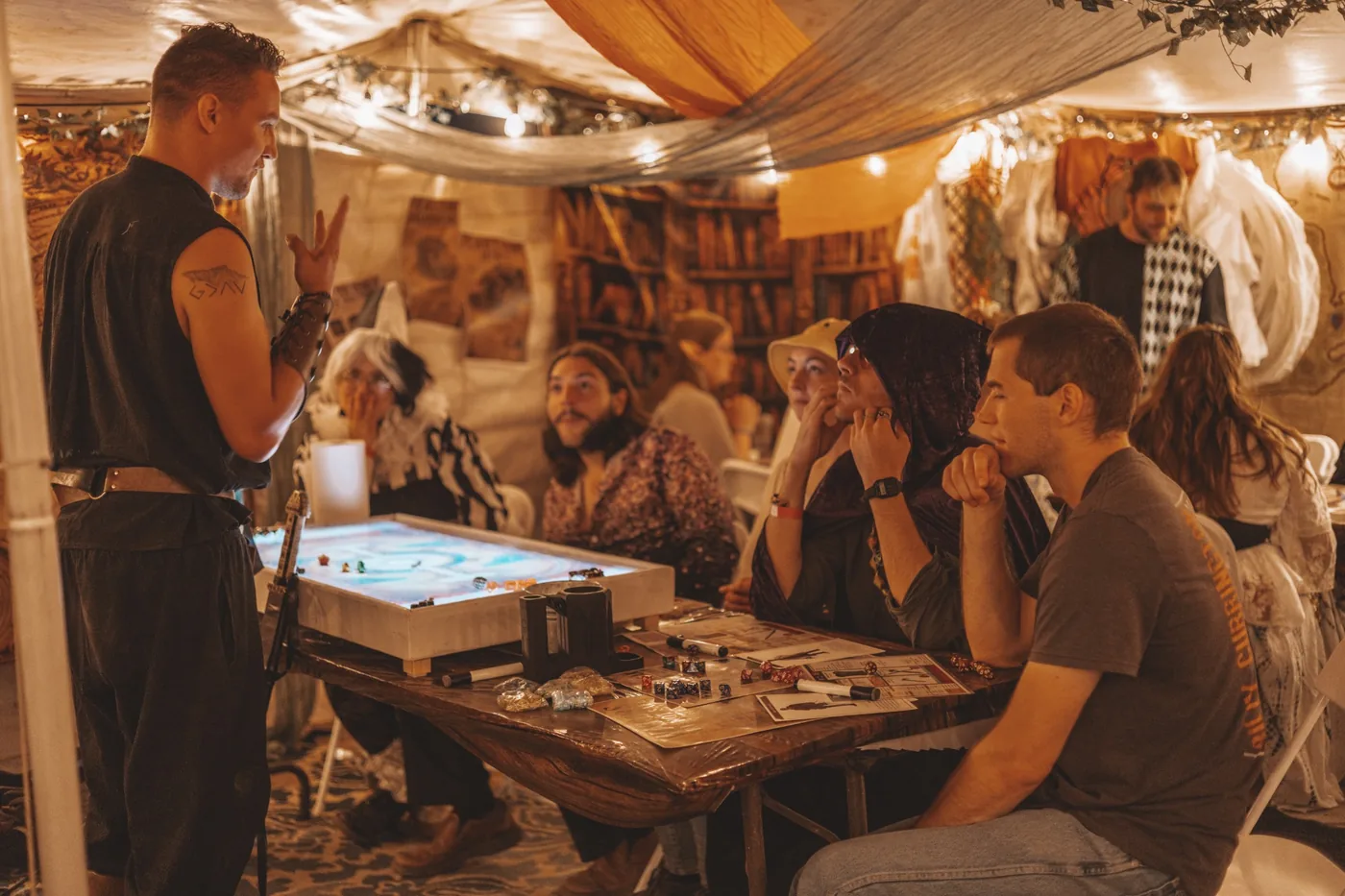 A costumed storyteller leading a group around a tabletop game in a warmly decorated event tent